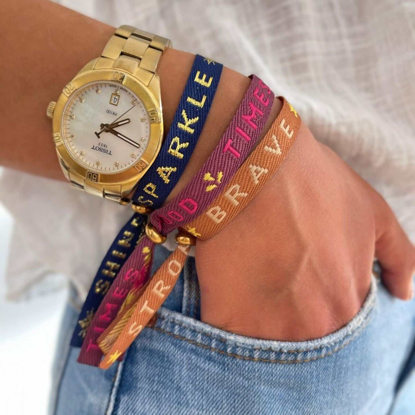 Bunte Statement-Armbänder am Handgelenk, gepaart mit einer goldenen Uhr, zeigen Worte für Positivität und persönliches Wachstum.