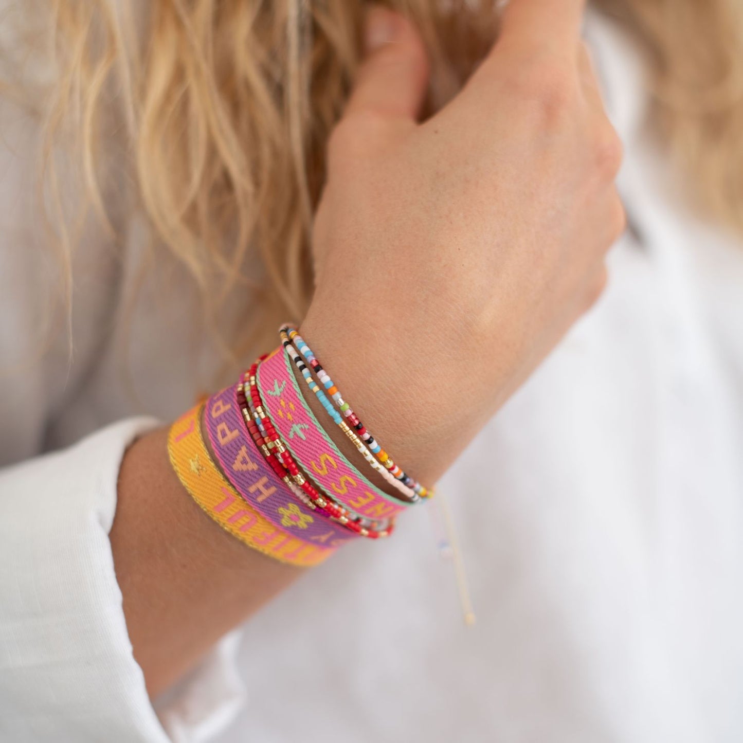 stoffarmbänder mit happy, life is beautiful, happiness und perlenarmbänder am arm einer frau mit weissen hemd