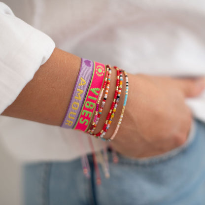 stoffarmband in neon pink mit good vibes und lila mit amour gewebt. mit perlenarmbändern am arm einer frau